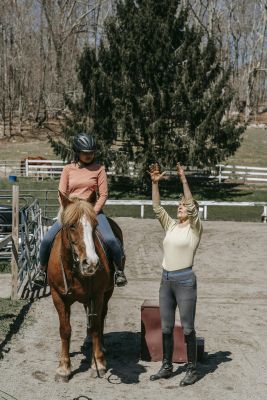 A trainer guides a woman on horseback outdoors during a sunny day lesson.